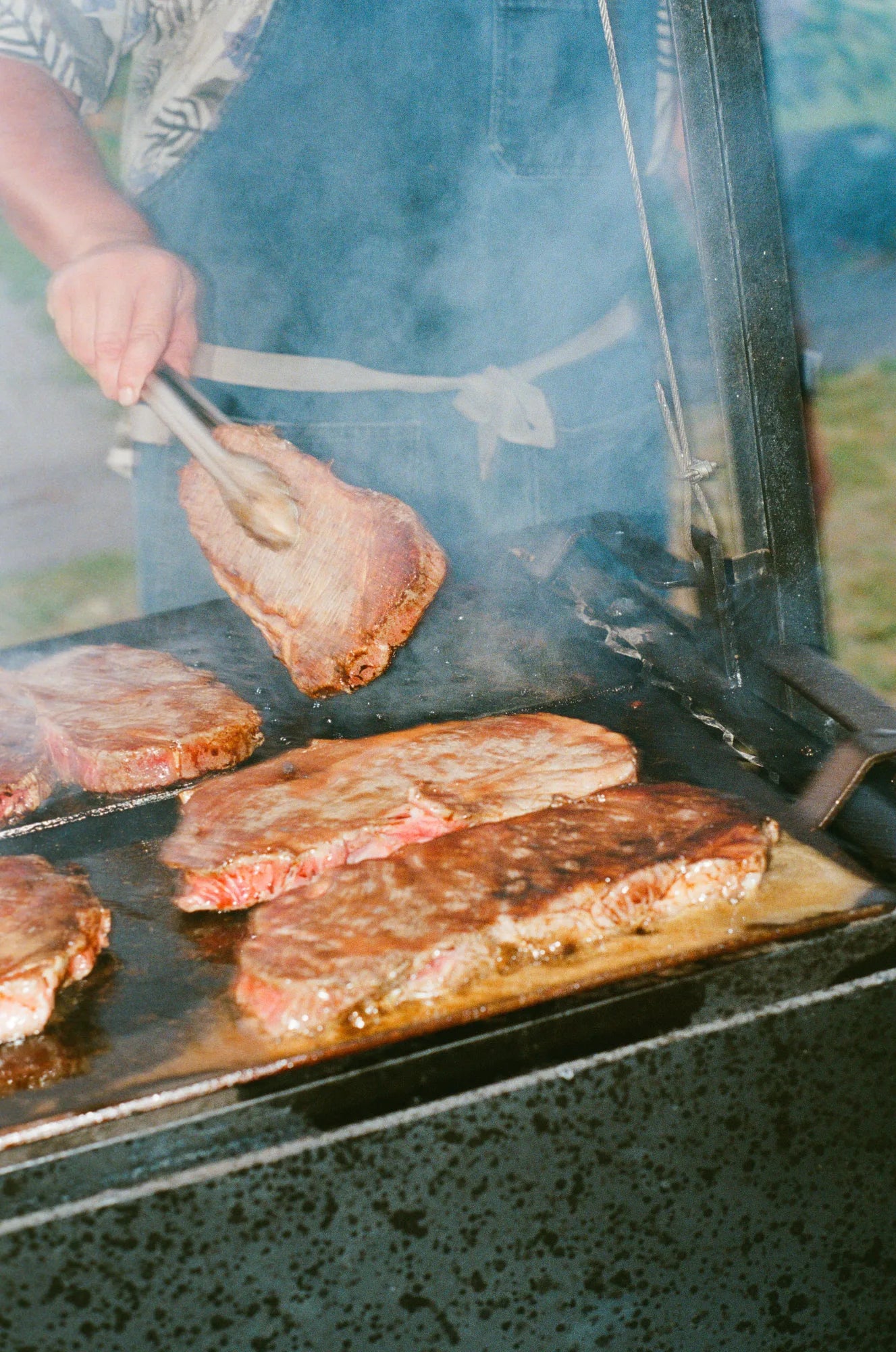 A cook in a denim apron flipping a Wagyu steak over billowing smoke on a live-fire grill