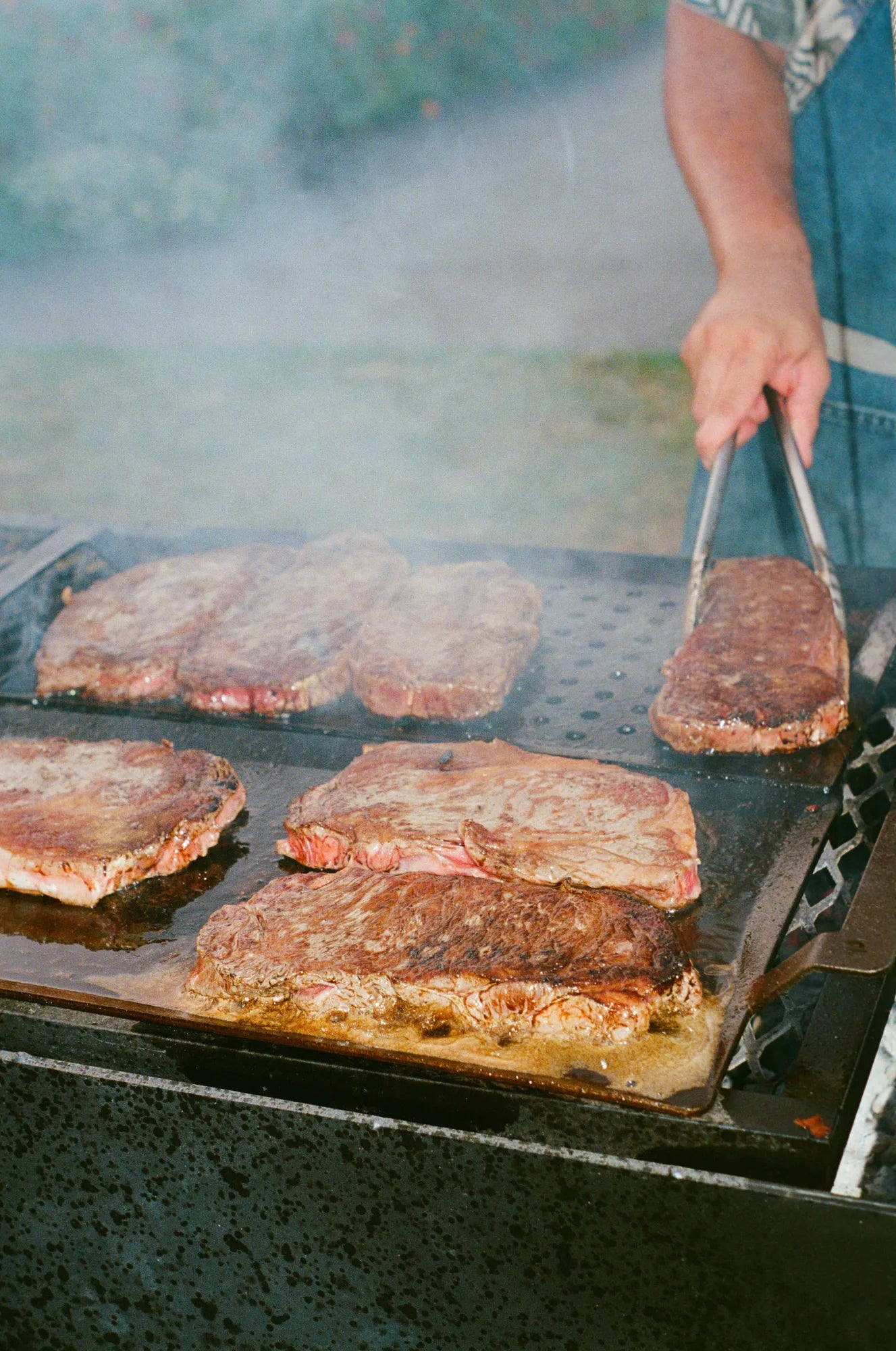 Wagyu steaks searing on a flat-top over open flame, a cook turning one with tongs