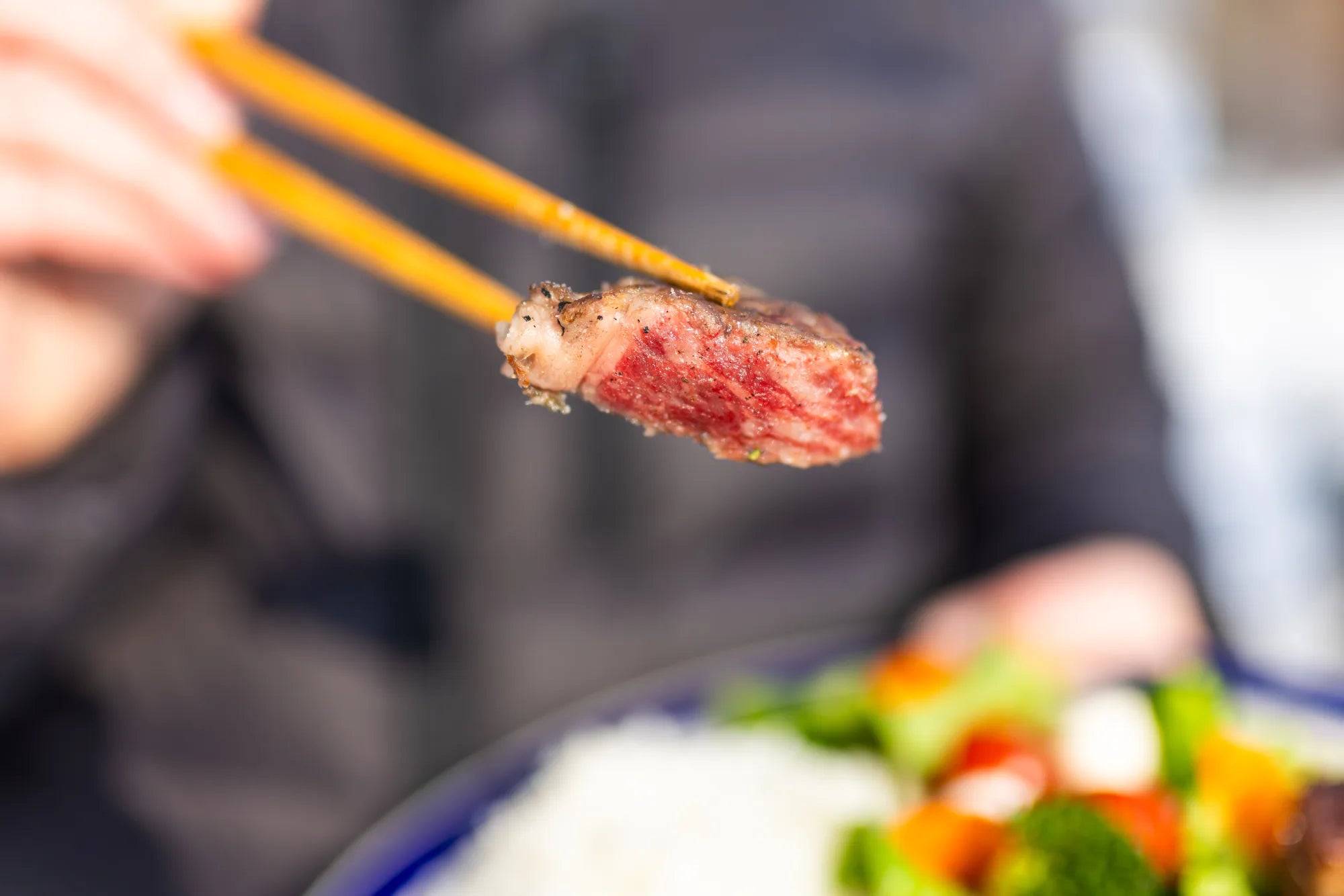 A cooked cube of Wagyu held aloft in chopsticks, showing the marbling in profile